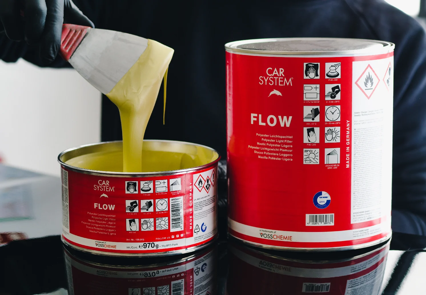 Two CARSYSTEM Flow Putty cans, one opened with a spatula lifting smooth, flowing putty.