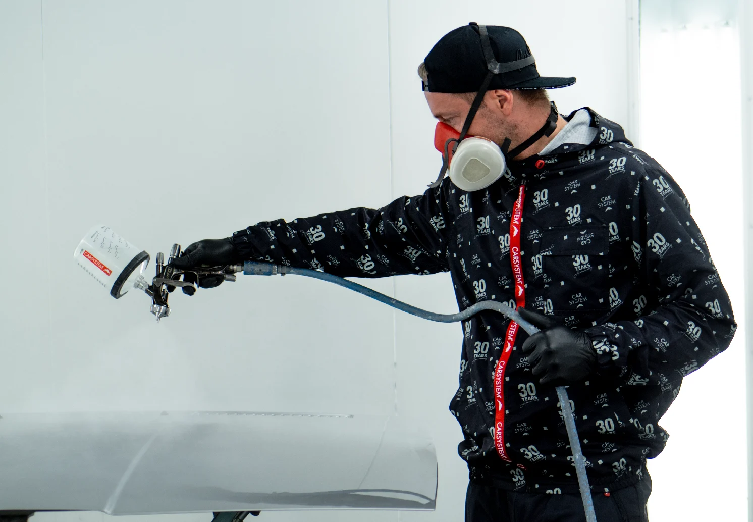 Technician wearing a CARSYSTEM anniversary coverall spraying a car panel with a paint spray gun.