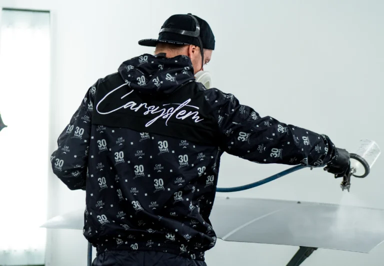 Technician wearing a CARSYSTEM anniversary coverall spraying a car panel with a paint spray gun.